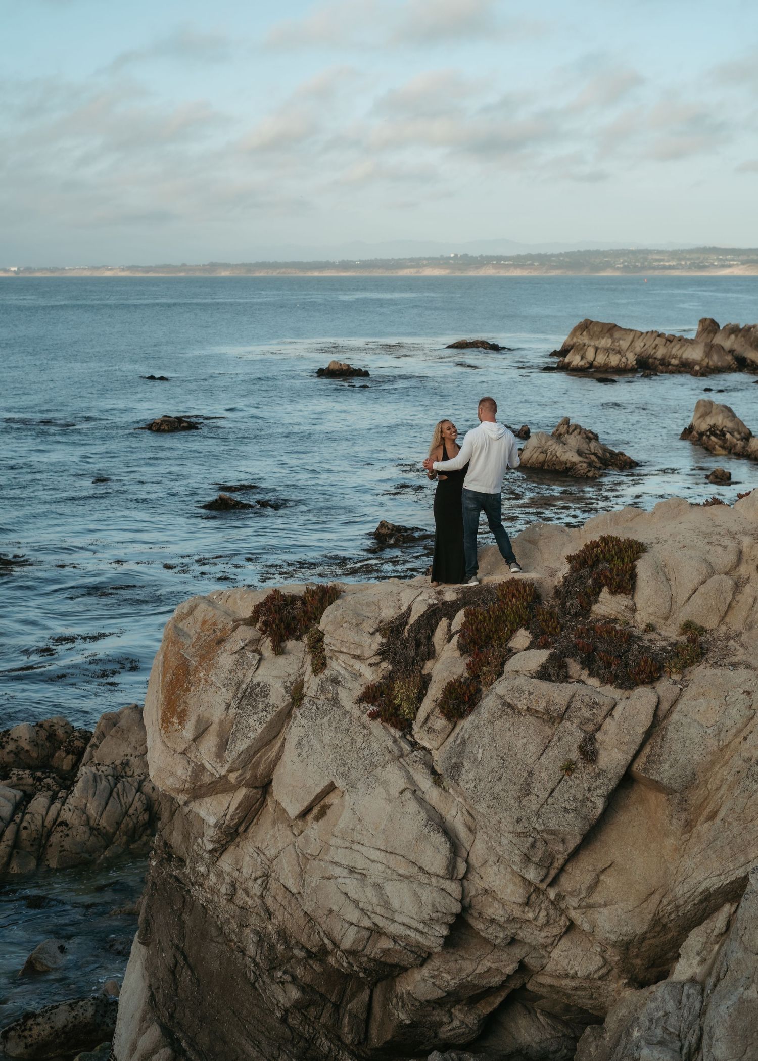 Four sequential shots of figures sitting on rugged coastal rocks overlooking the Pacific Ocean during a cloudy day.
