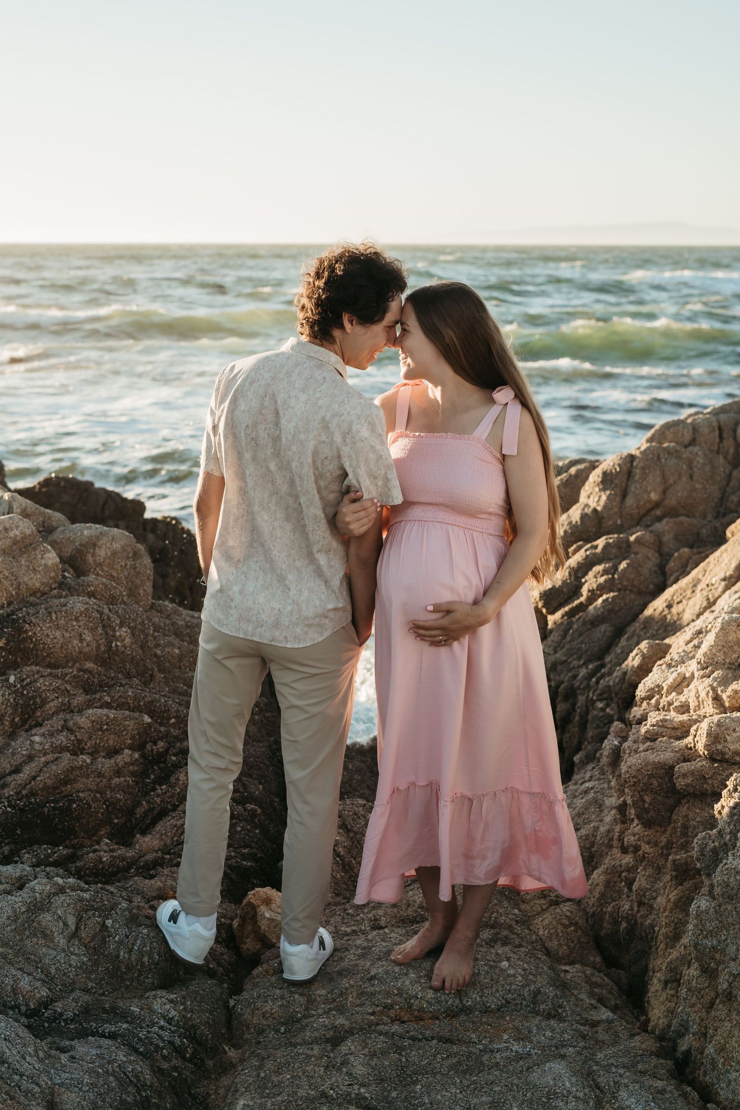 A couple embraces on rocky beach at sunset as gentle waves crash behind them.