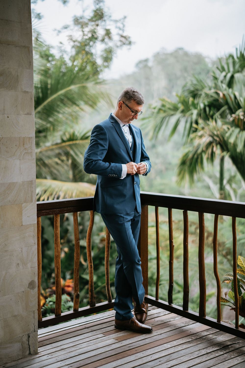 A groom in navy suit checks his watch on a balcony overlooking tropical scenery.
