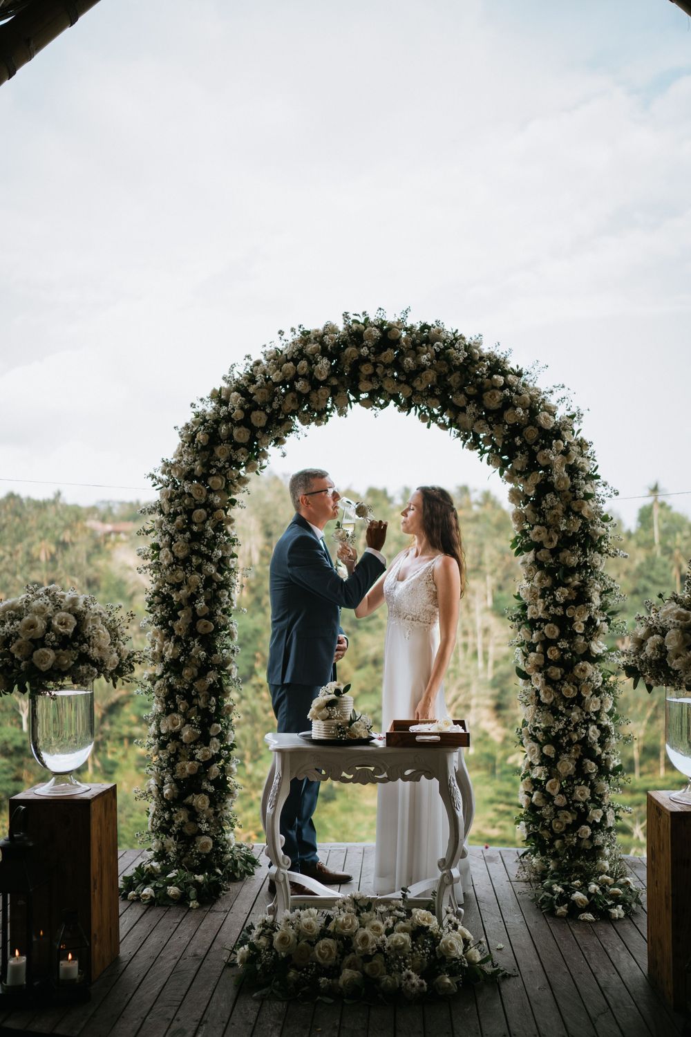 Newlyweds share intimate moments under an elegant white floral arch on a wooden deck with scenic views.