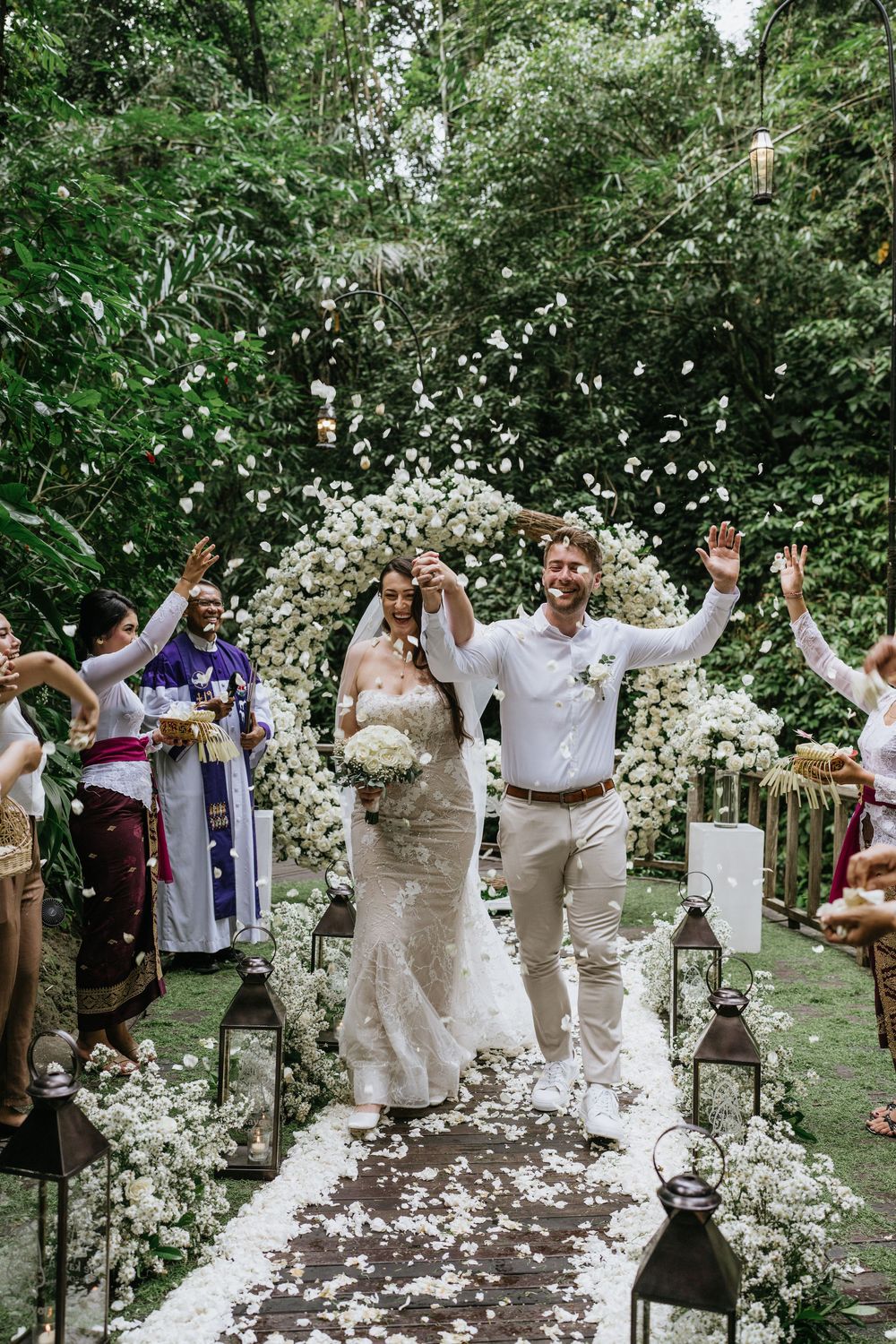Wedding guests throw white flower petals during an outdoor garden ceremony celebration.