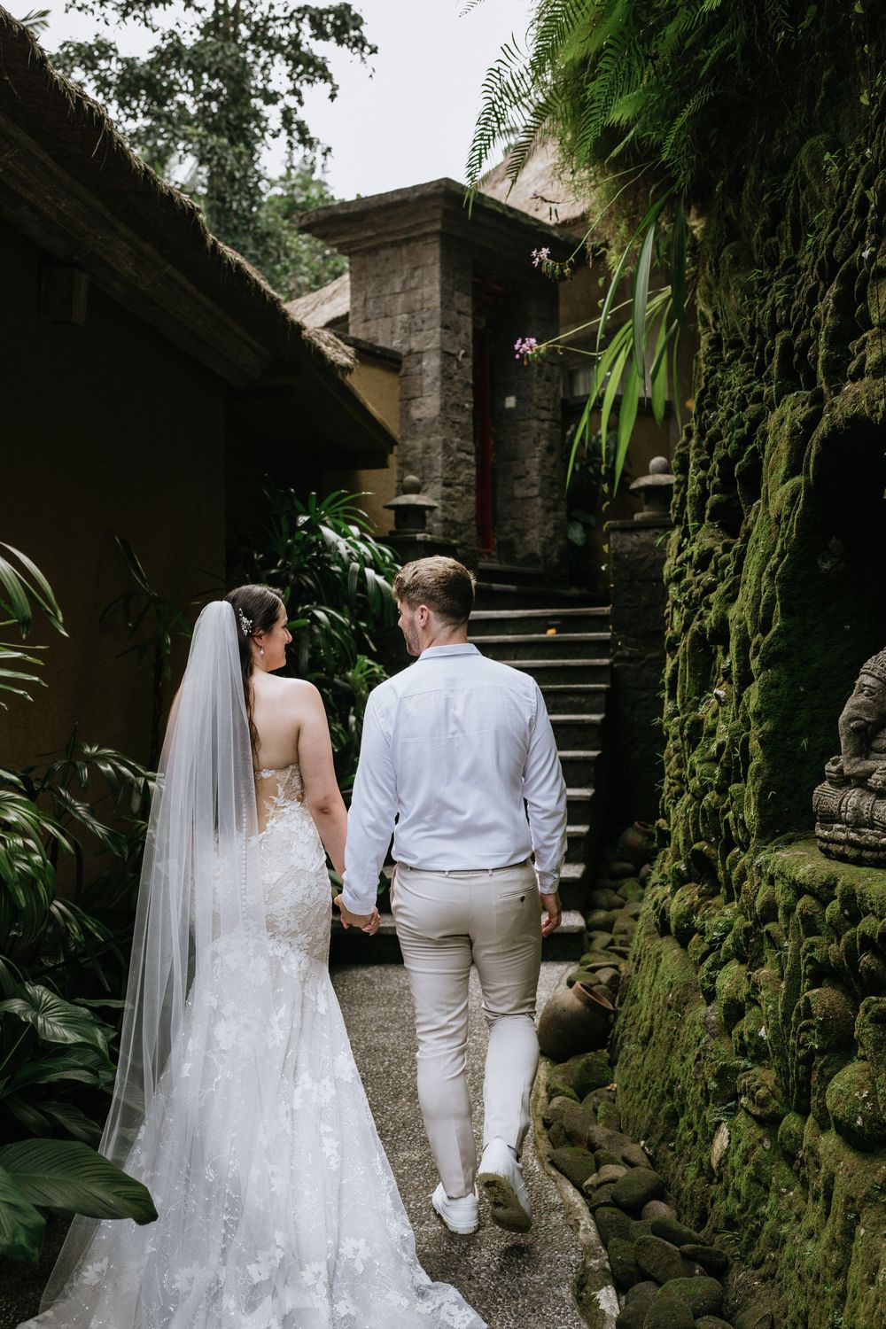 A couple in wedding attire walks along a stone path surrounded by tropical greenery and moss-covered walls.