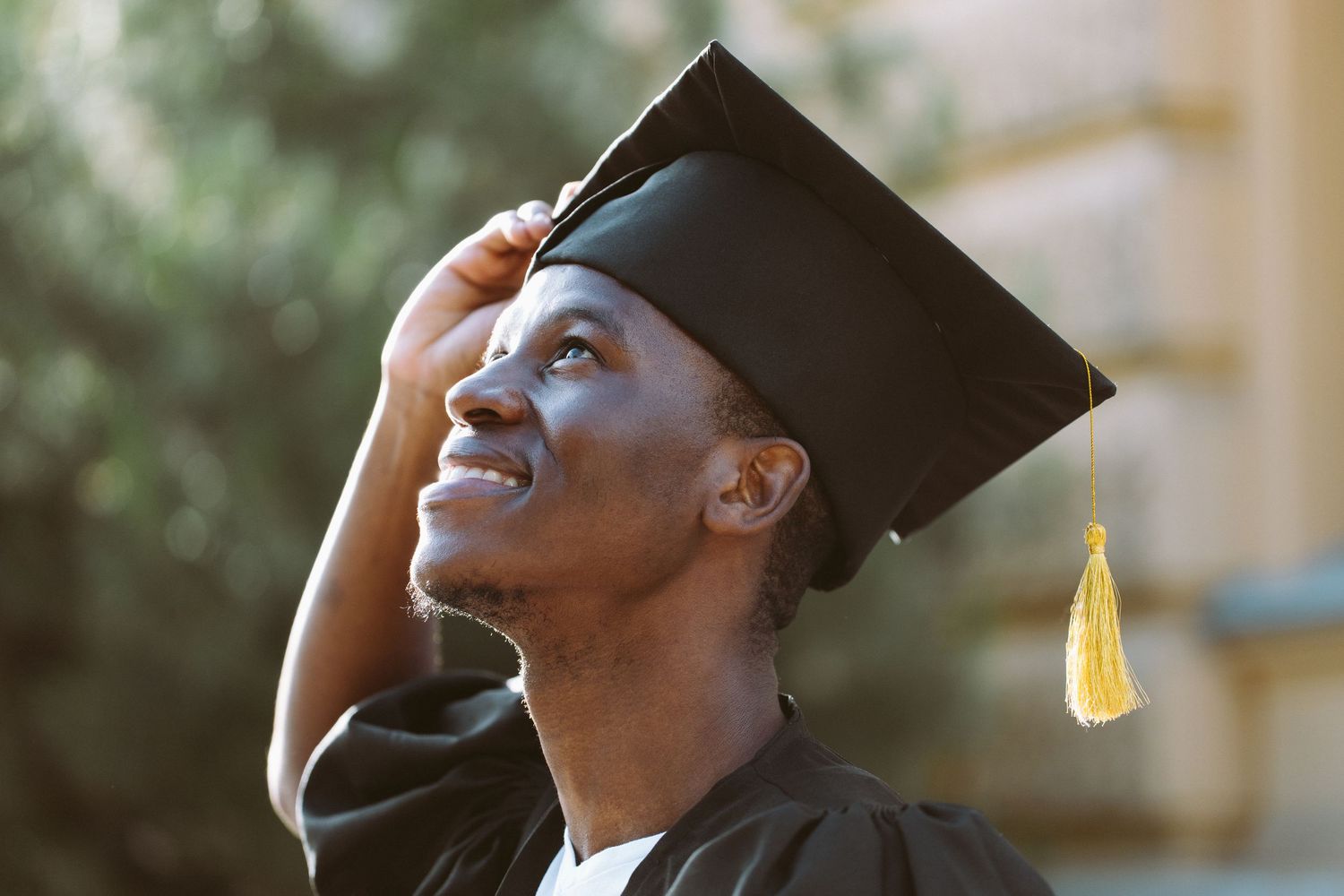 Graduate in cap and gown looking up and smiling with sunlight creating warm glow in background.