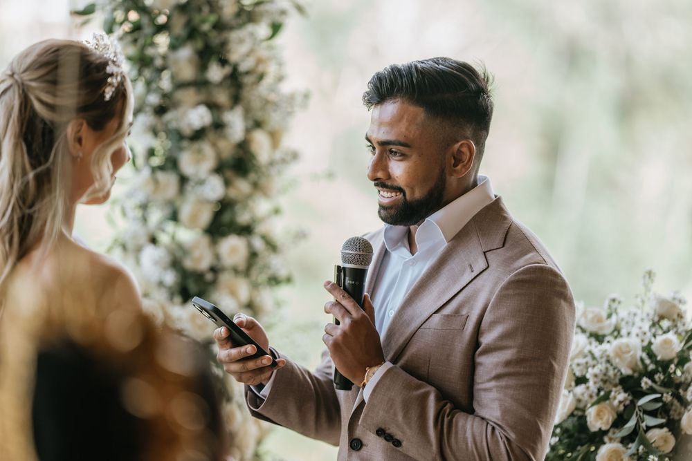An emotional wedding ceremony moment under a floral arch as vows are exchanged with a microphone.