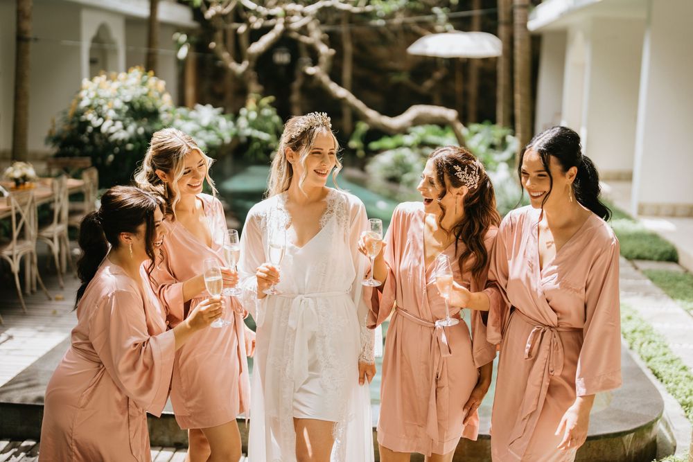 Bridesmaids in pink robes gather around the bride in white during wedding preparations.