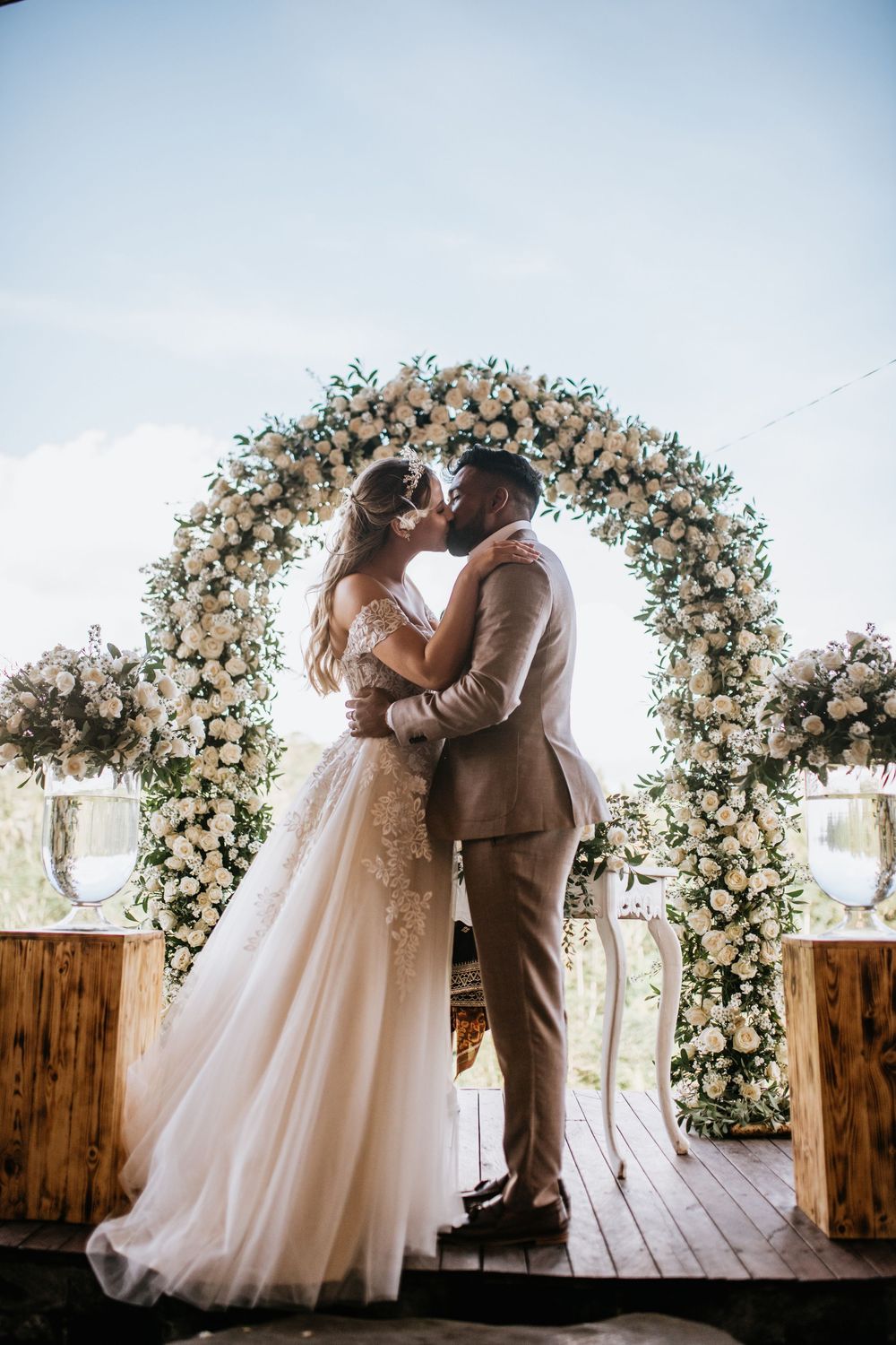 Romantic kiss under elaborate white floral arch during outdoor wedding ceremony.