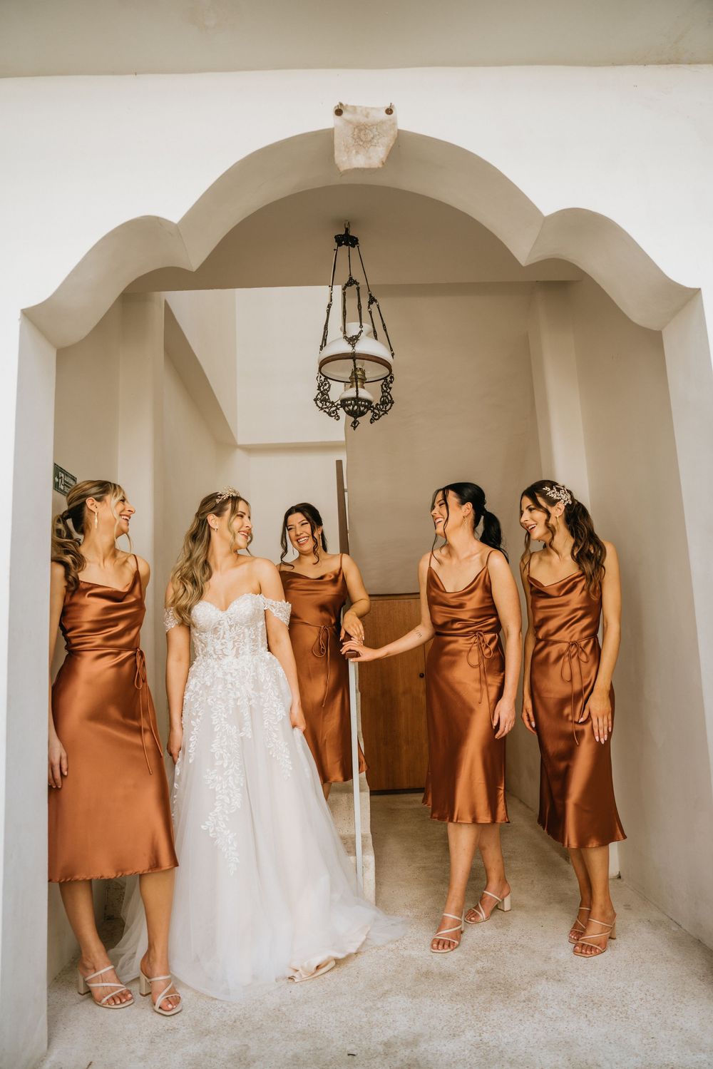 Bridal party in brown silk dresses and white gown pose playfully in arched hallway.