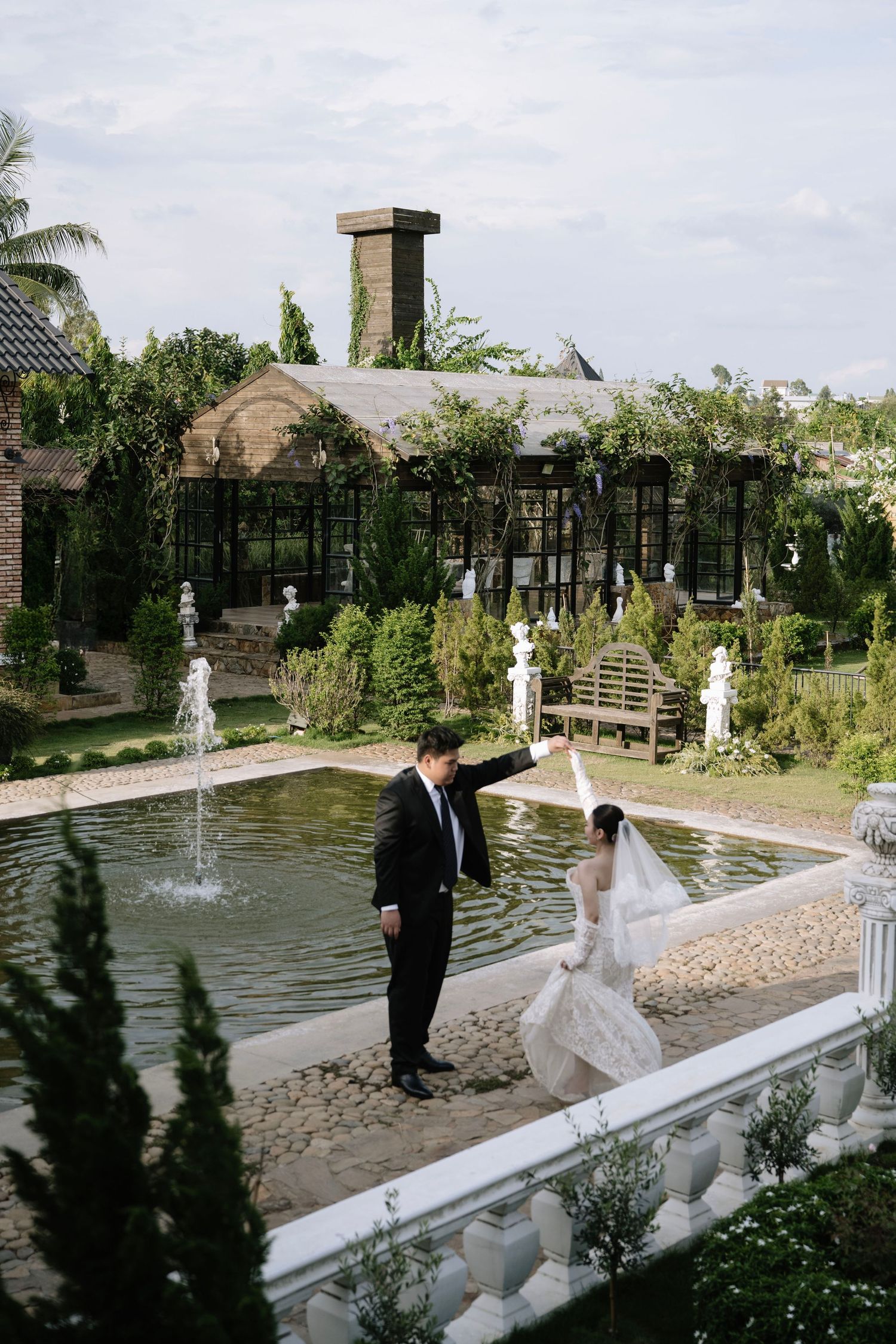 Bride and groom pose by garden fountain at lush wedding venue with greenery.