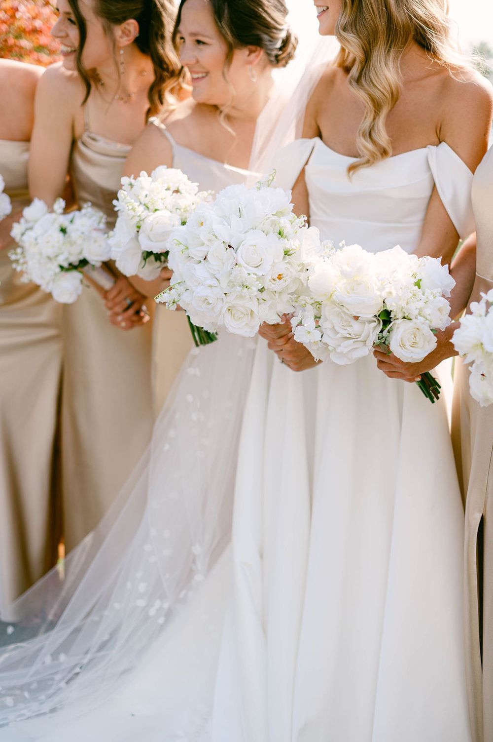 Bridesmaids in elegant dresses holding white floral bouquets stand together in warm sunlight.