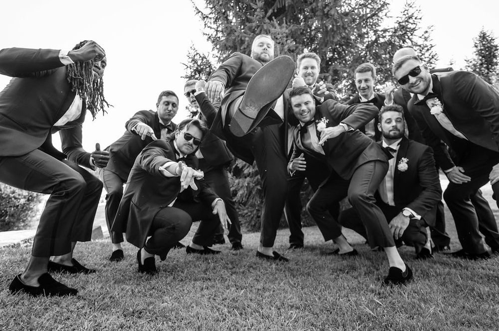 Groomsmen in black suits kneeling and posing dramatically in a line on grass outdoors.