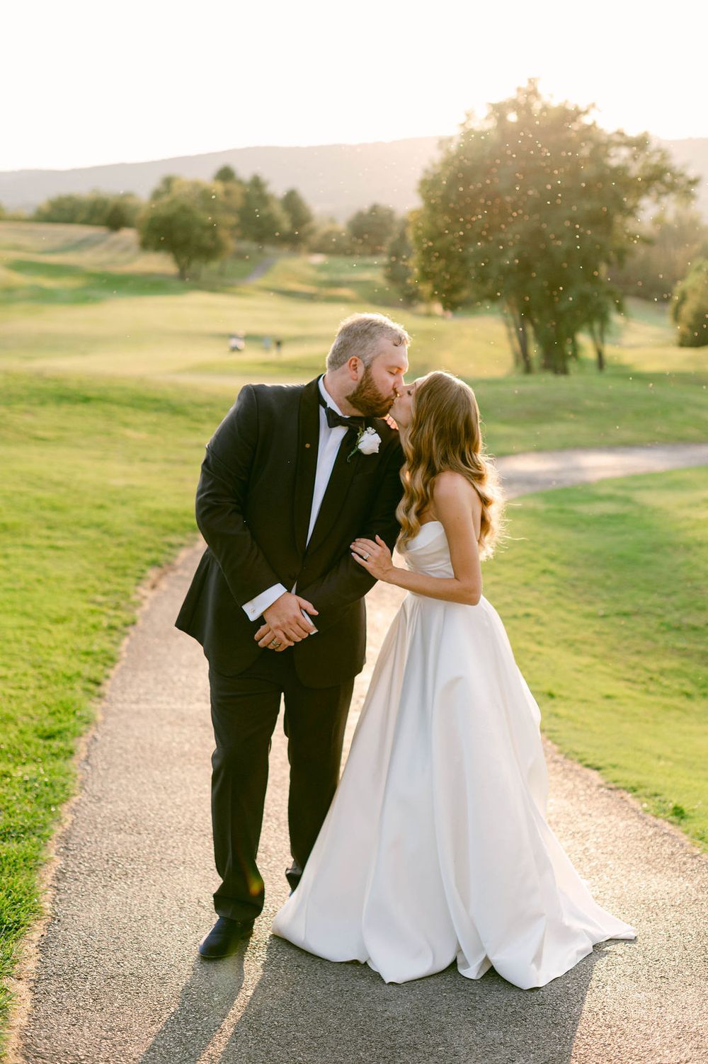 Newlyweds share an intimate moment on a path overlooking a scenic golf course at sunset.