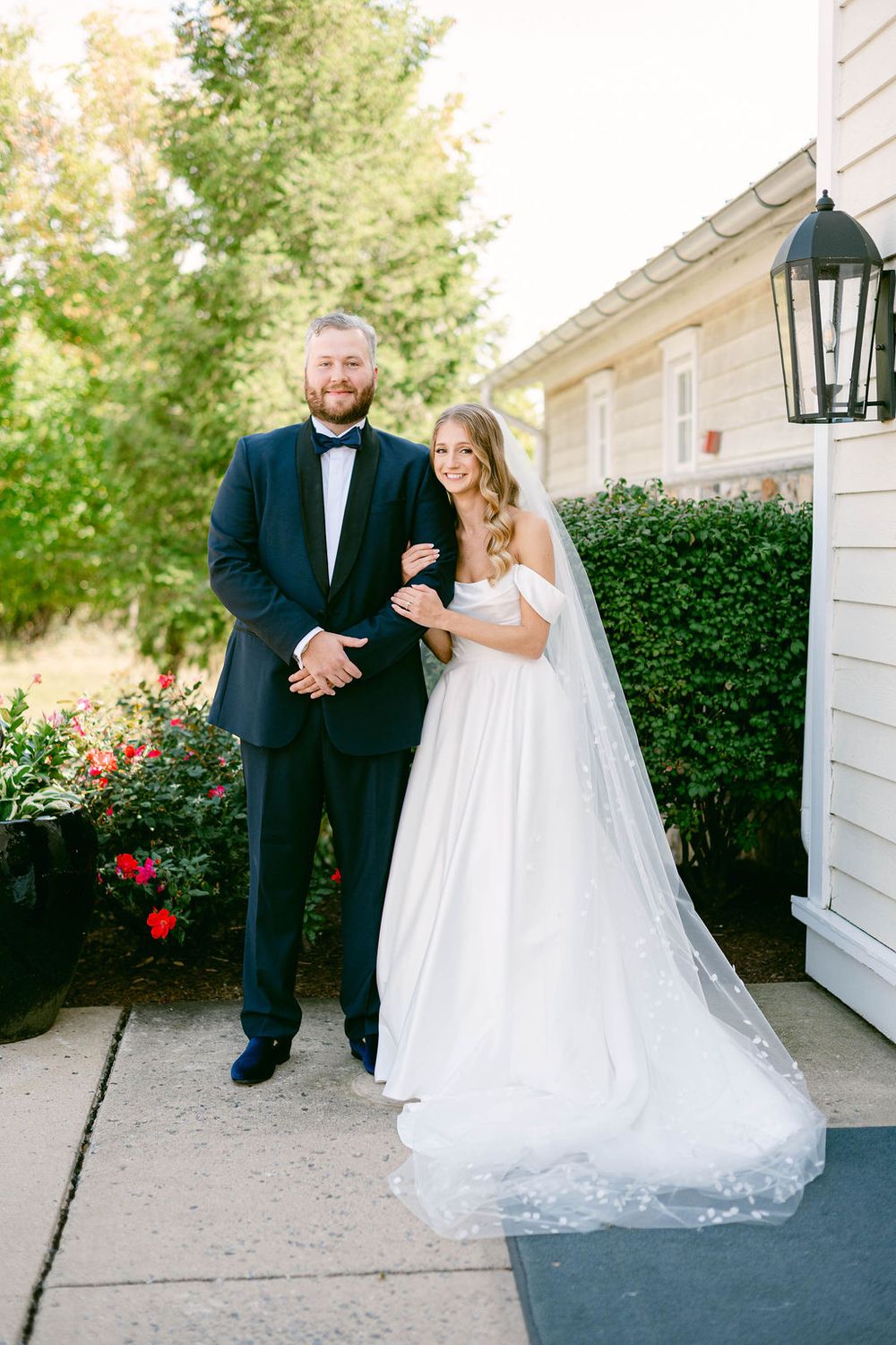 Wedding couple walks along a garden path beside a white building with blooming red roses.