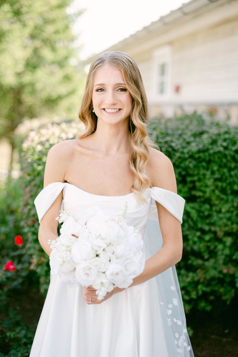 Bride in elegant off-shoulder white dress holds classic white bouquet in garden setting.