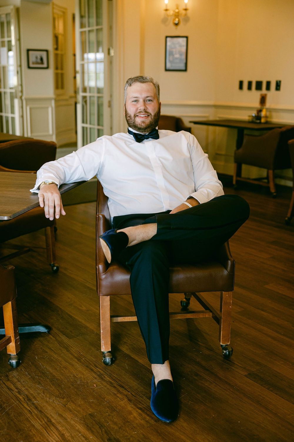 Groom relaxes in brown leather chair while taking casual getting-ready photos in warm-lit room.