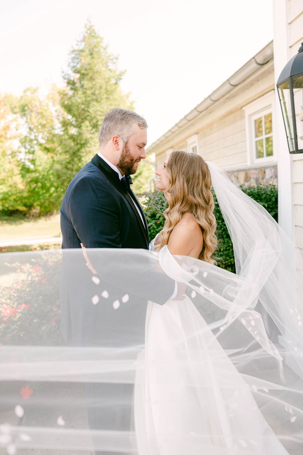 Romantic couple in wedding attire share an intimate moment outside with flowing veil in sunlight.