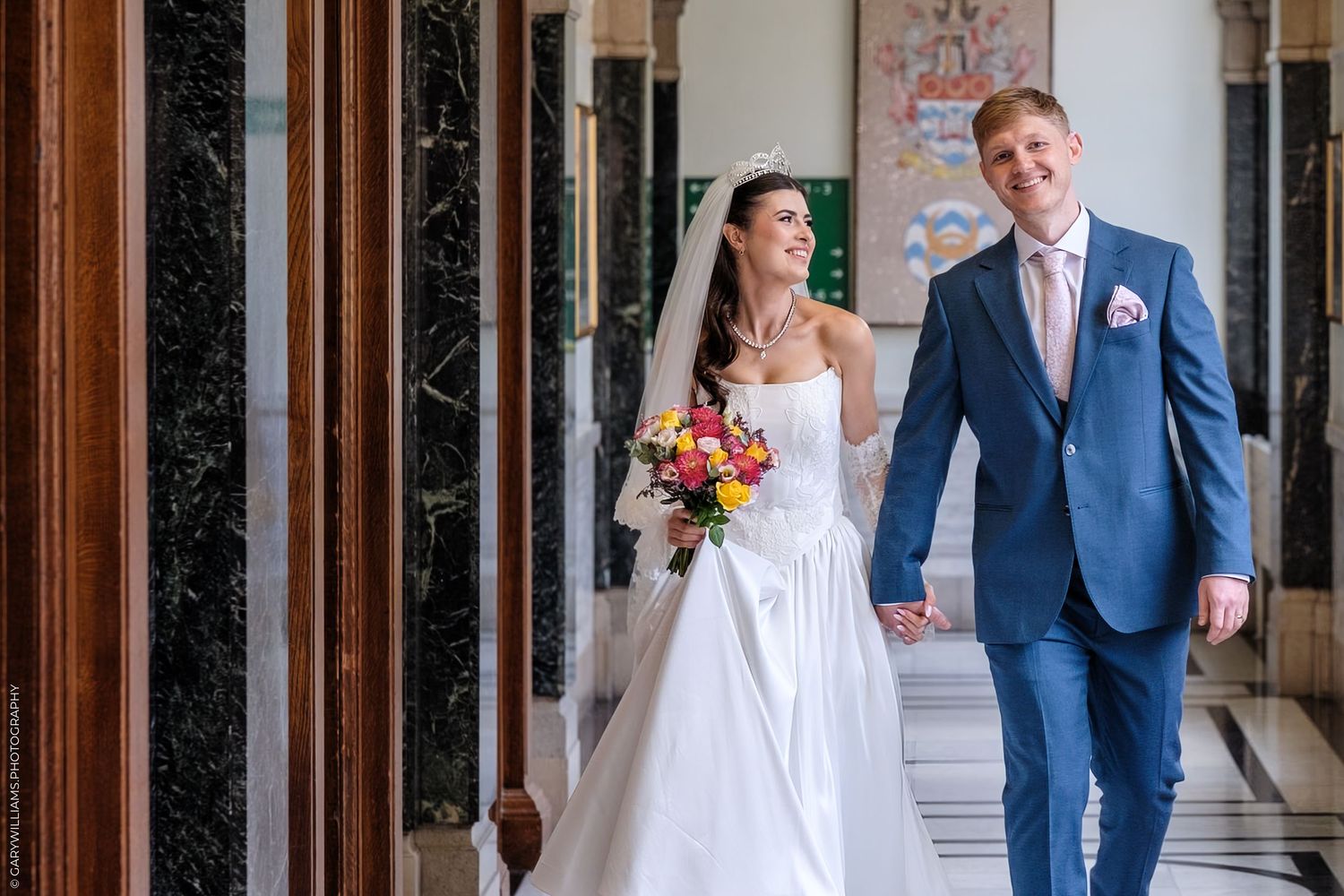 A bride and groom walk through a grand columned corridor with stone pillars and sunlight streaming in.