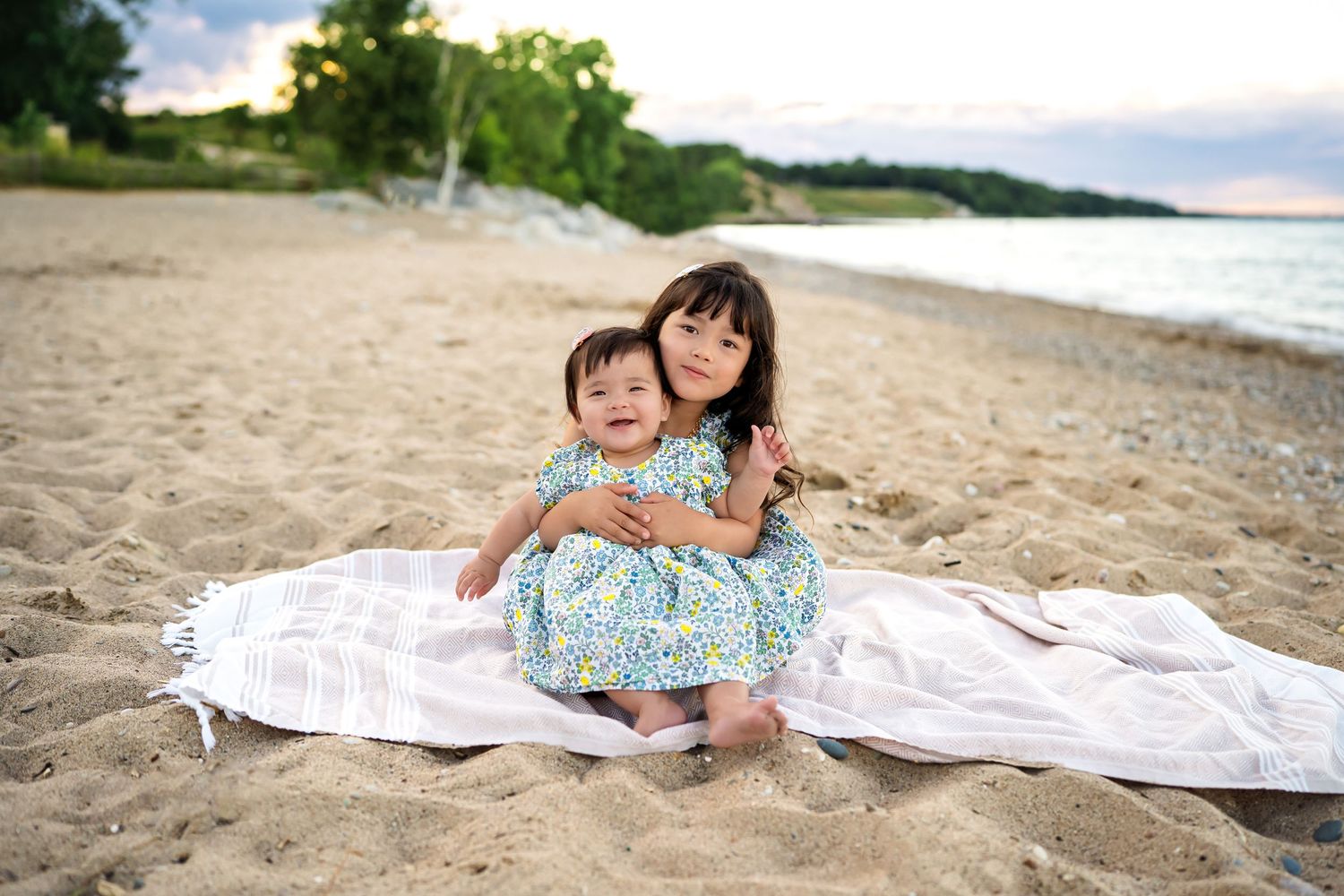 Two people sit together on a white blanket at a scenic beach with rolling waves and lush greenery in the background.