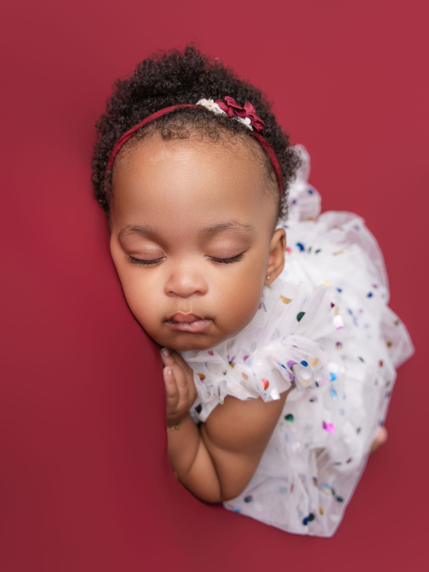 Sleeping newborn in white tulle dress with colorful dots on deep red background.