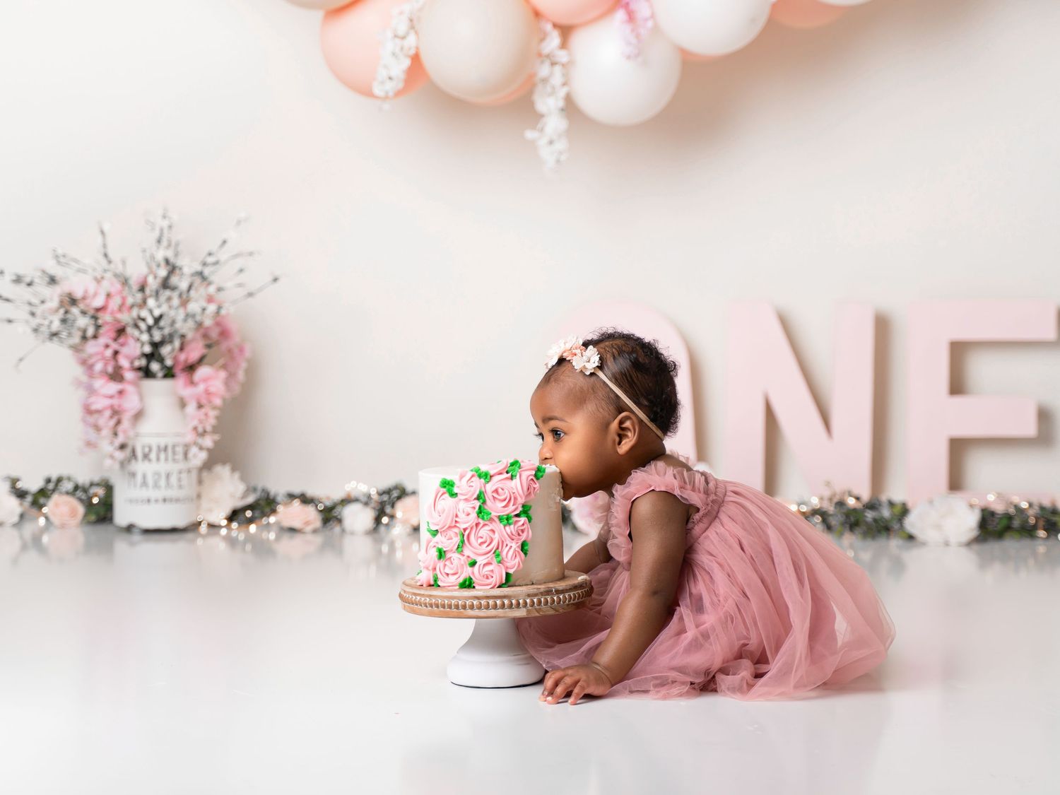Baby girl in pink tutu dress leaning into birthday cake on white backdrop with balloons.