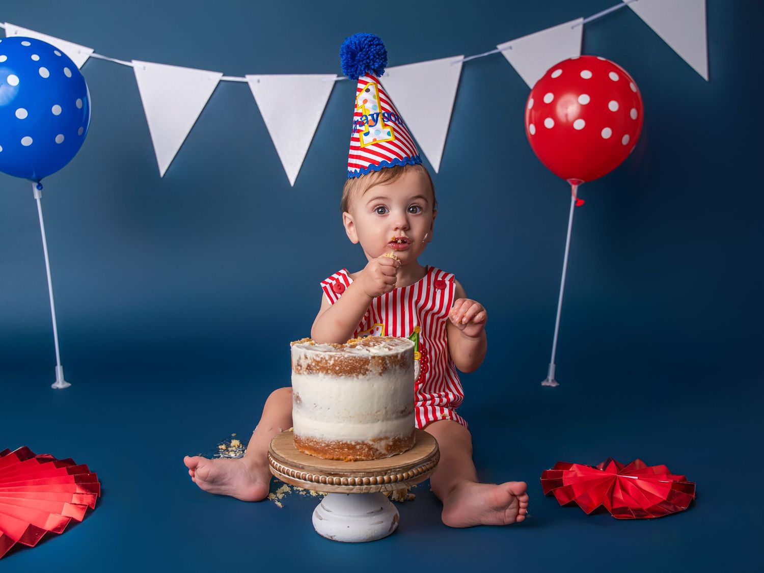 Baby in patriotic outfit eating first birthday cake with red and blue balloons on blue backdrop.