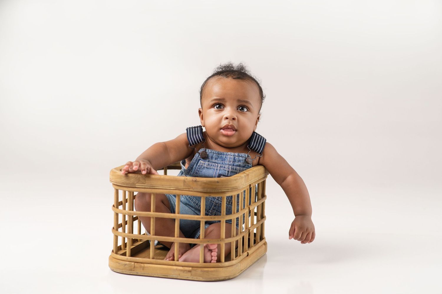 Cute baby boy in denim overalls peeks over edge of a wicker basket on white backdrop.