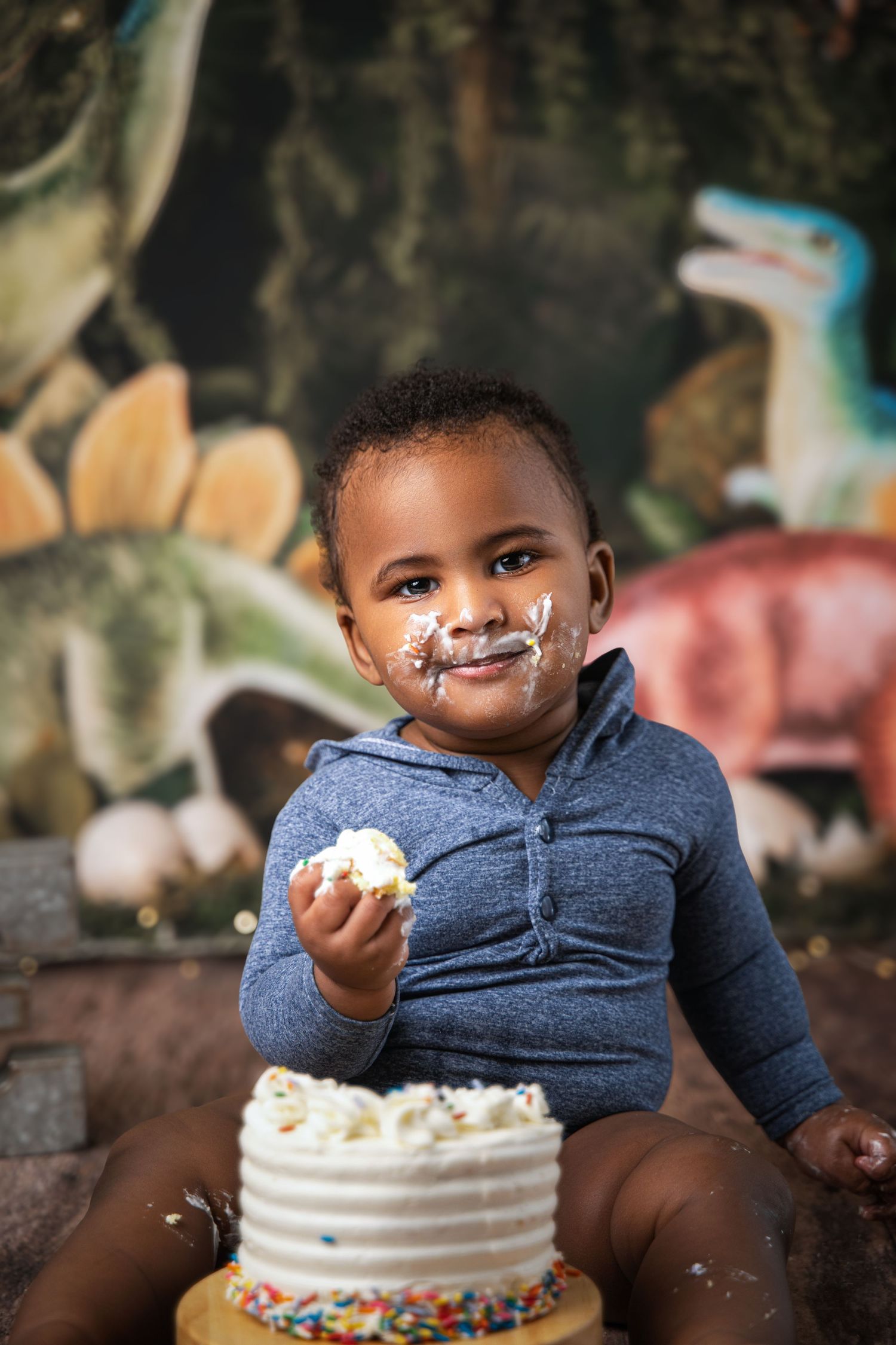 Toddler with cake frosting on face holds piece of smash cake at dinosaur themed first birthday photo session.