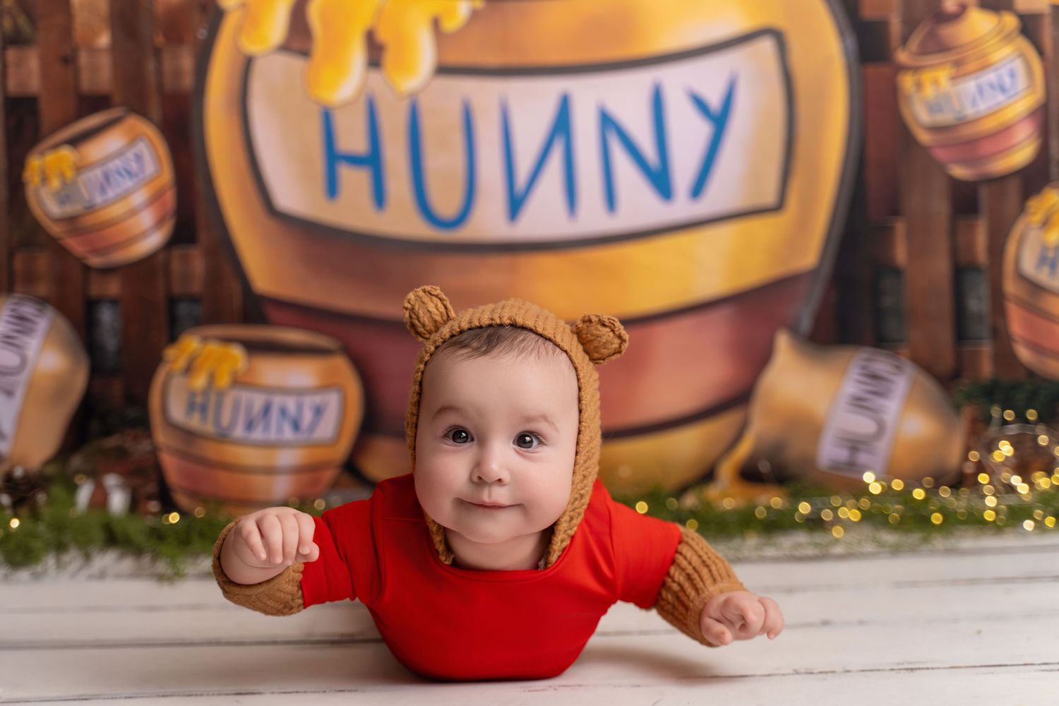 Adorable baby in bear bonnet and red onesie poses in front of Winnie the Pooh Hunny backdrop.