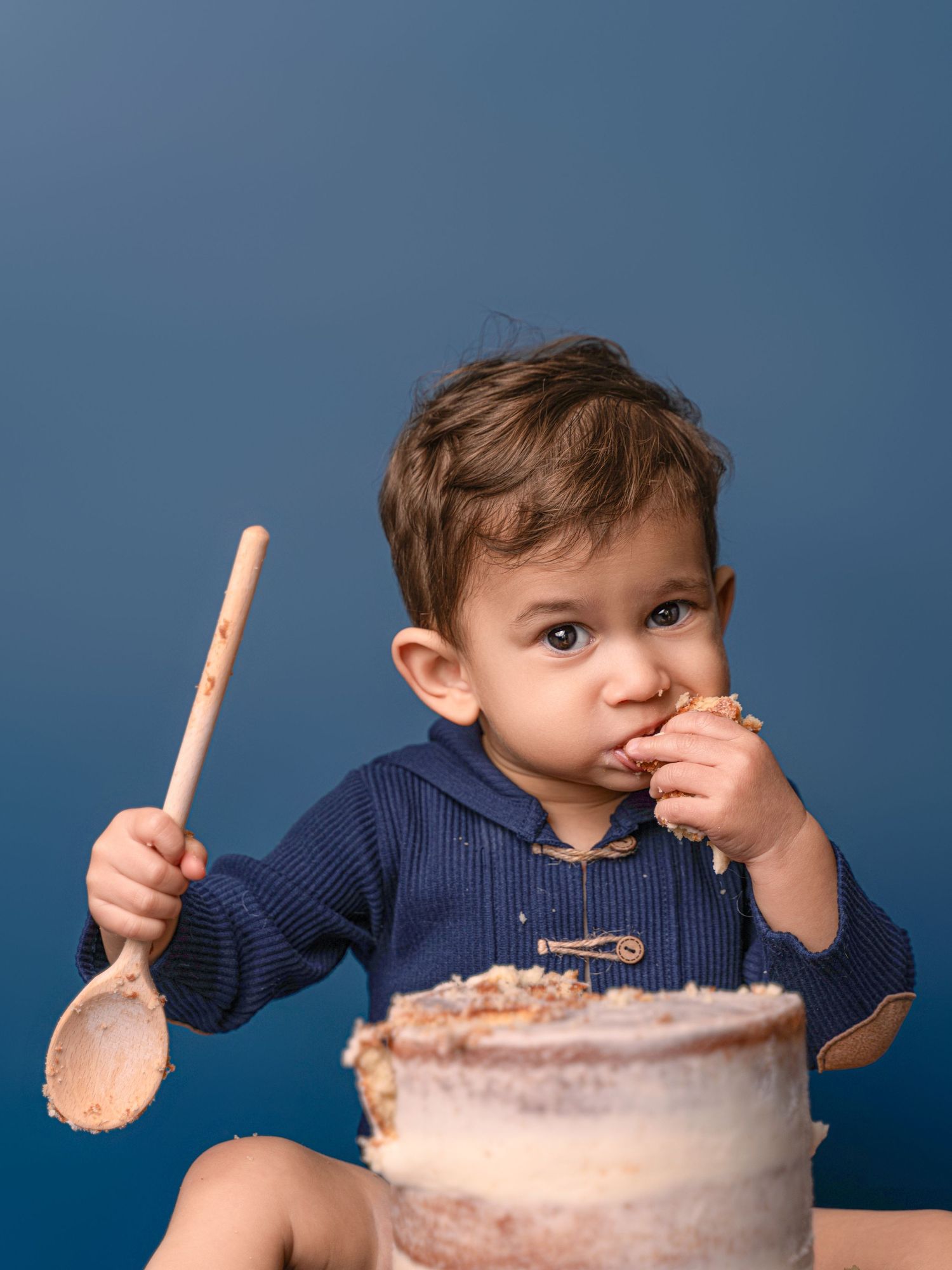 Curious toddler boy in navy hoodie eats cake while holding a wooden spoon on blue backdrop.
