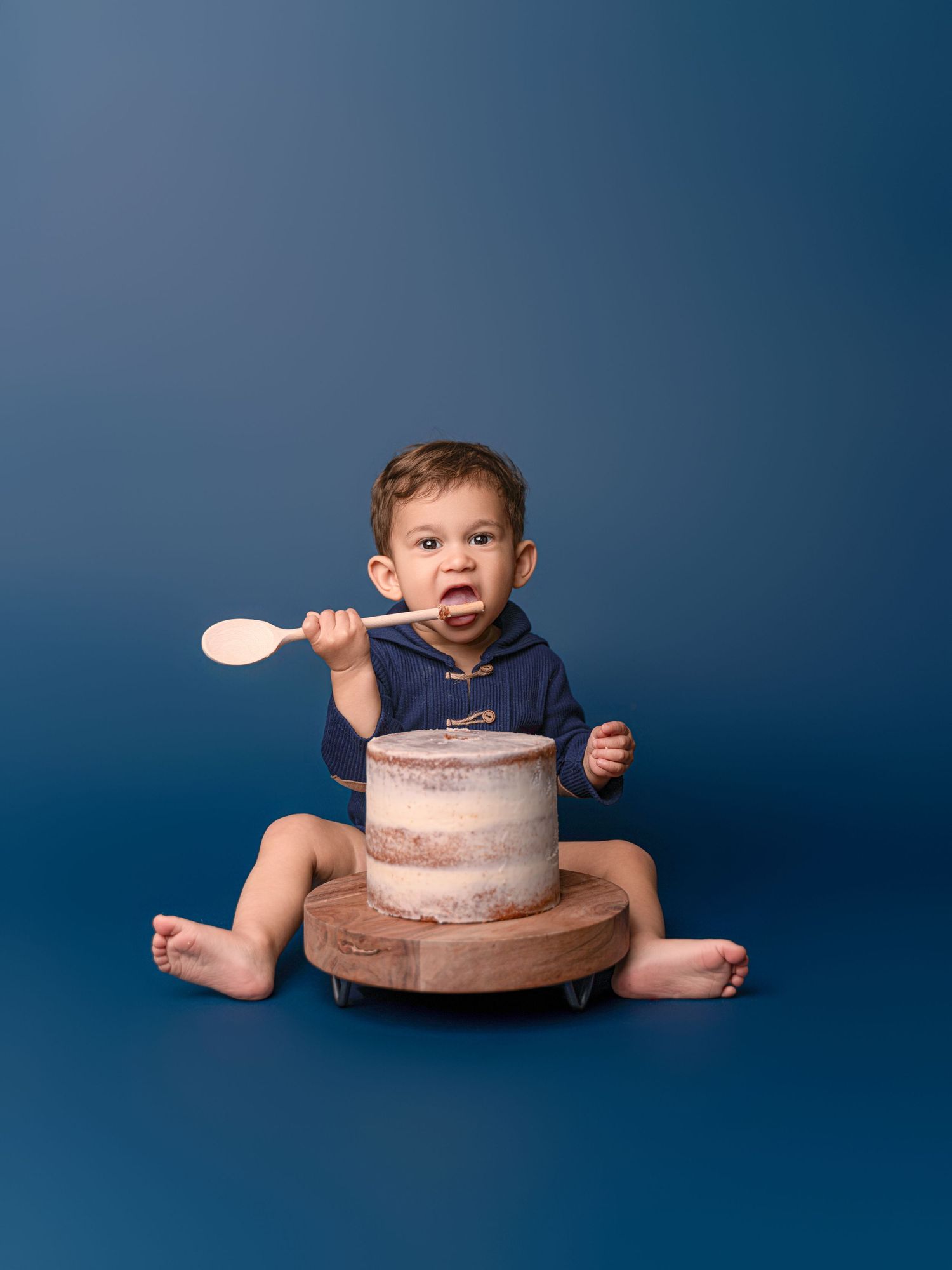 Baby boy in navy romper sits with wooden spoon beside a rustic cake on blue backdrop.