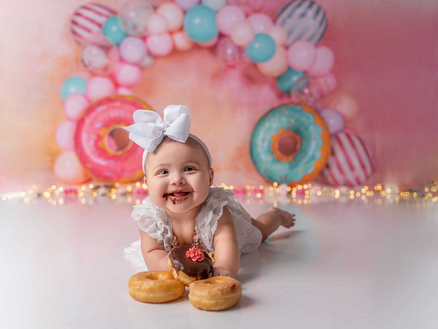 Happy baby girl in white dress smiles surrounded by donuts and colorful balloon arch backdrop.