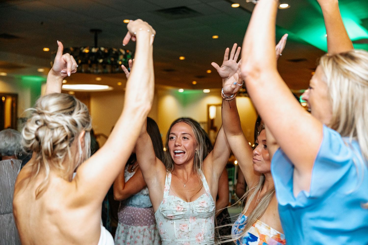 People dancing and celebrating with raised arms in a festive indoor venue with colorful lighting.