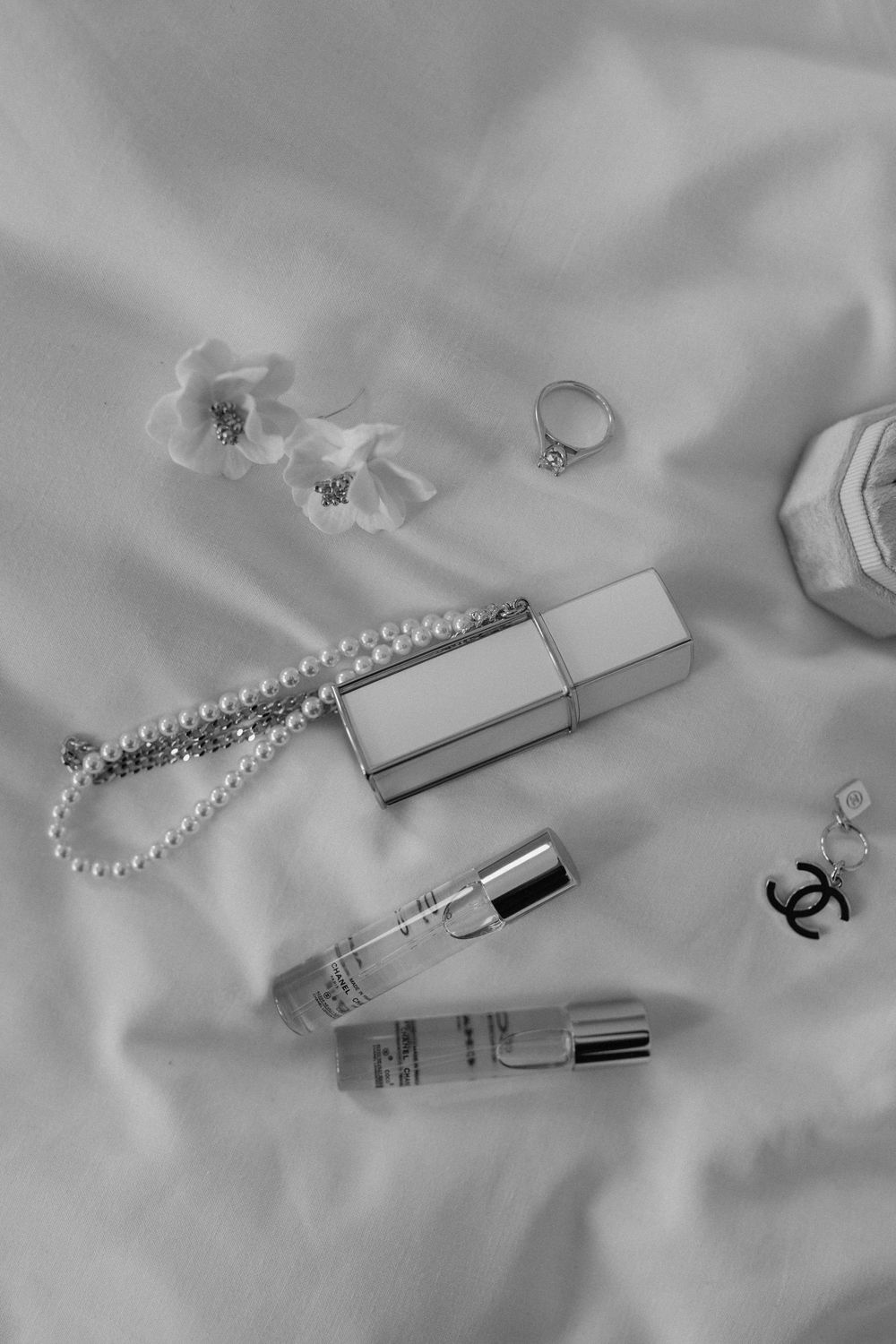 Black and white photo of beauty accessories including pearl necklace, rings, perfume bottles and Chanel jewellery on silk fabric.