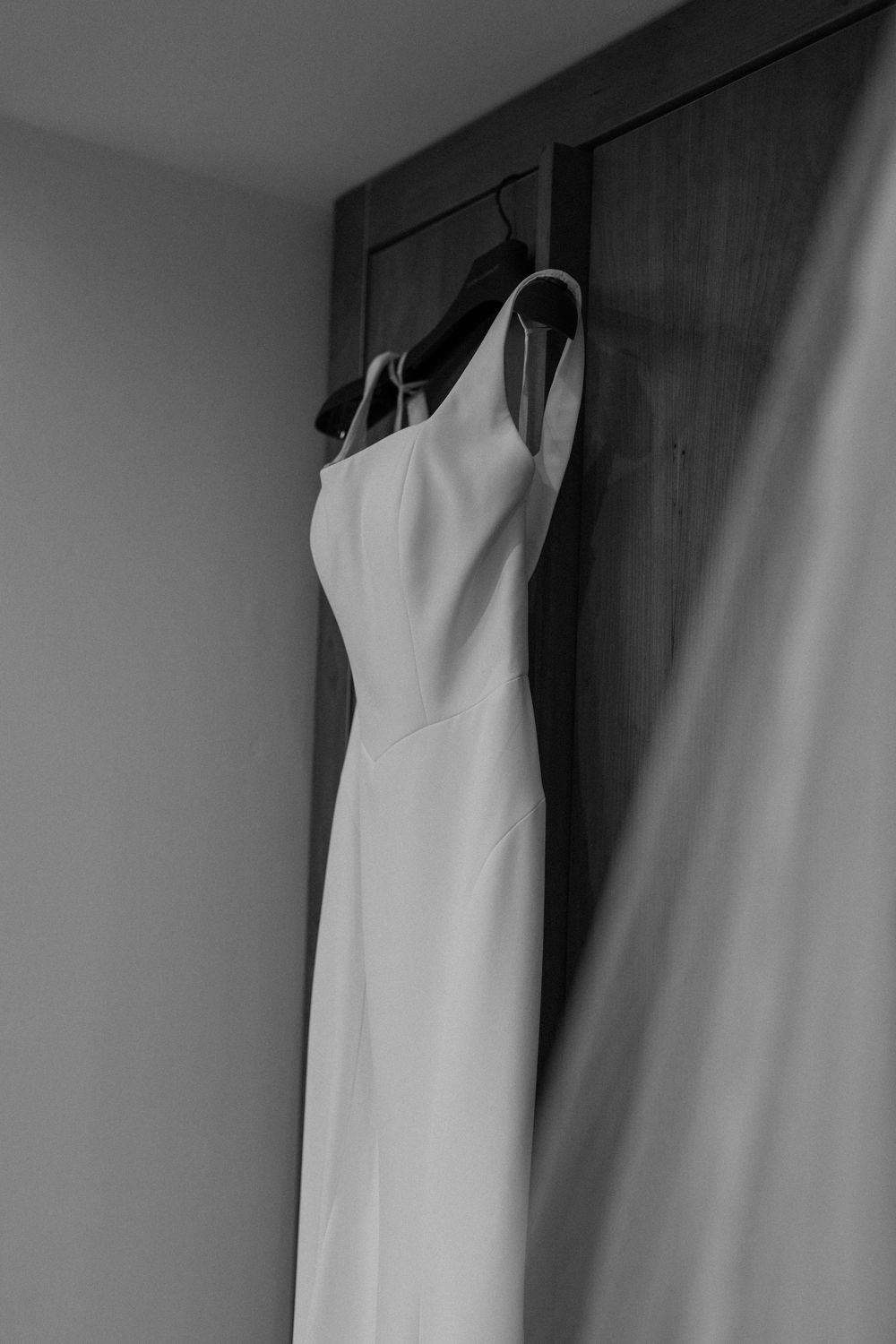 A minimalist white wedding dress hangs on a wooden door in a monochrome composition.
