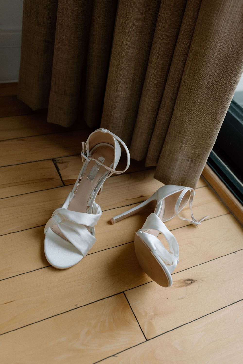 White strappy high heel sandals are arranged in a sequence on light wooden flooring by curtains.