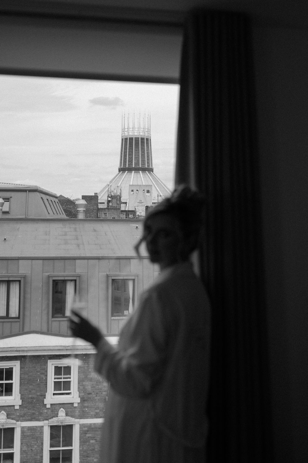 A silhouetted bride stands by a window overlooking Liverpool's Metropolitan Cathedral and urban rooftops in black and white.