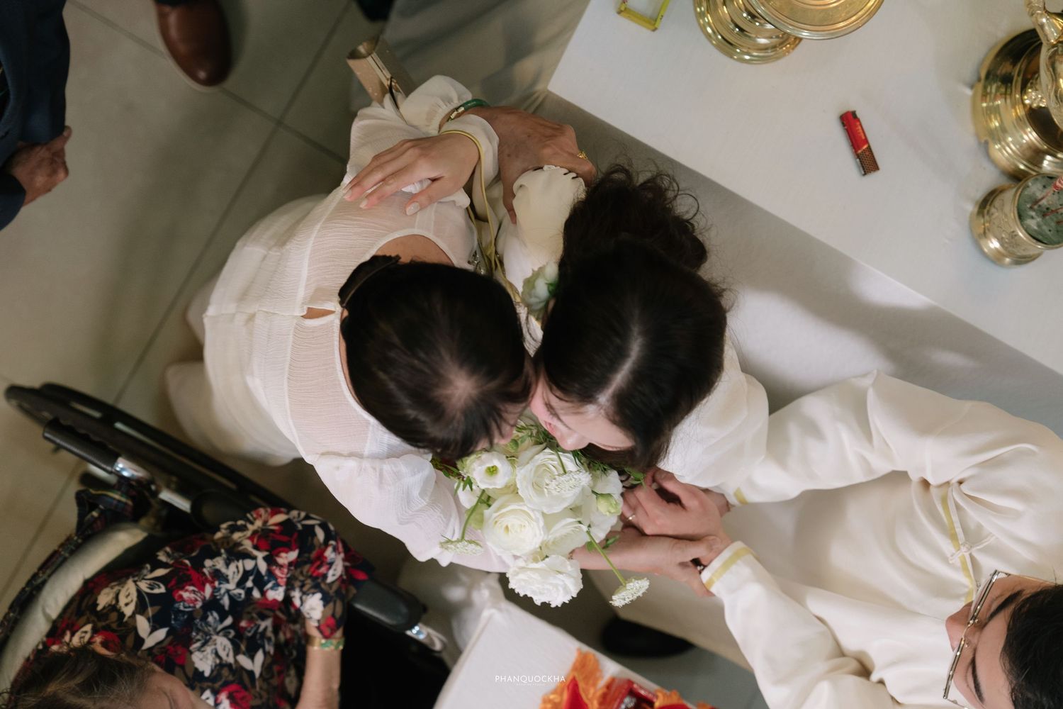 Overhead view of Vietnamese wedding couple in white ao dai holding white bouquet during traditional ceremony.