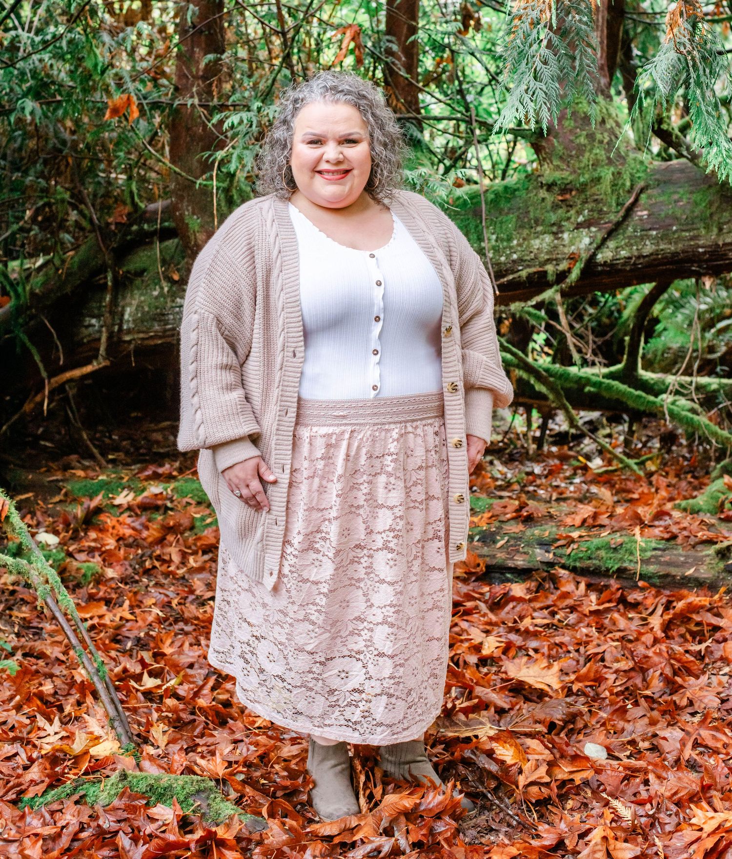 Person wearing beige cardigan over white dress stands among autumn foliage in wooded area.