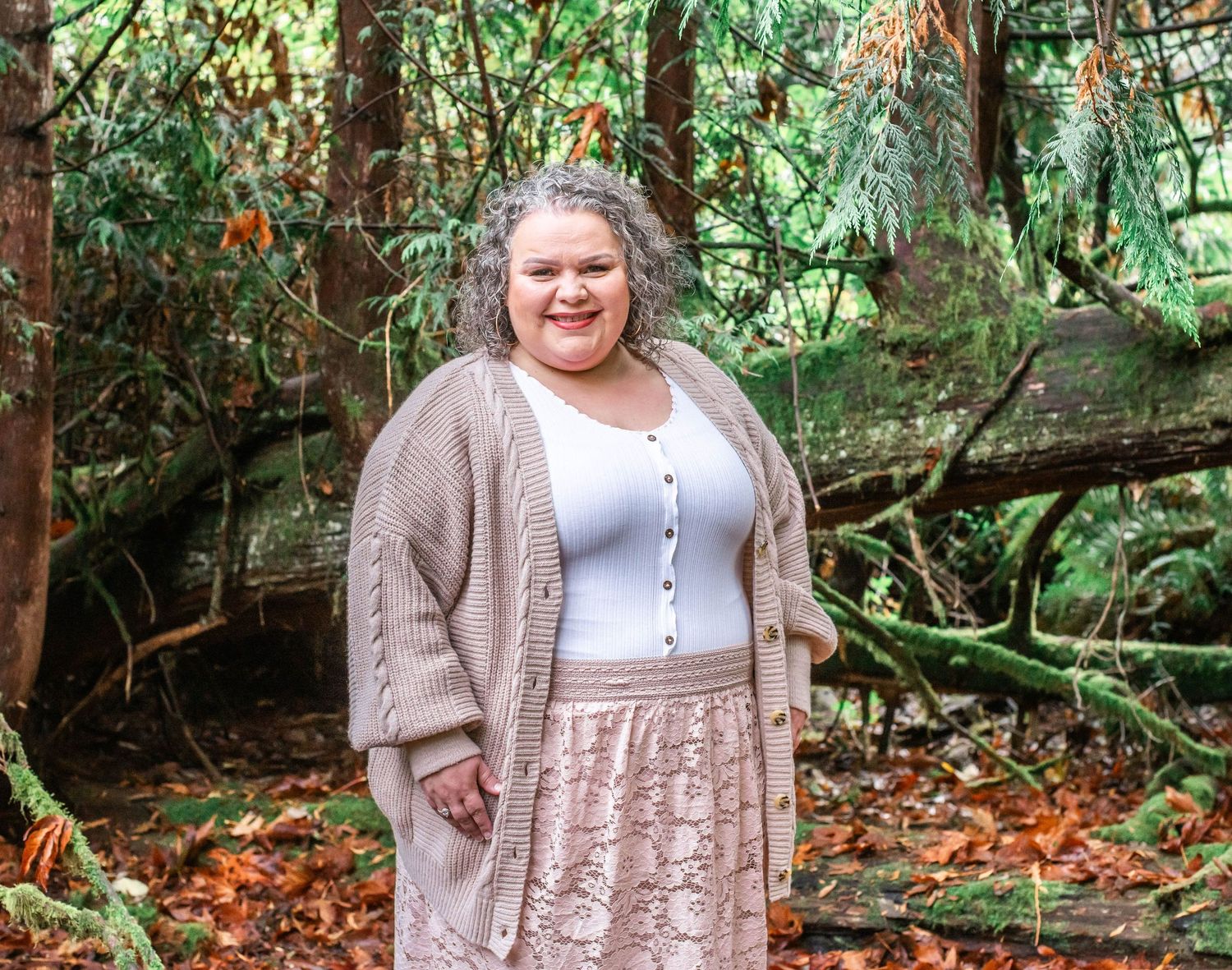 Person in beige cardigan and dress poses among fallen autumn leaves in forest setting.