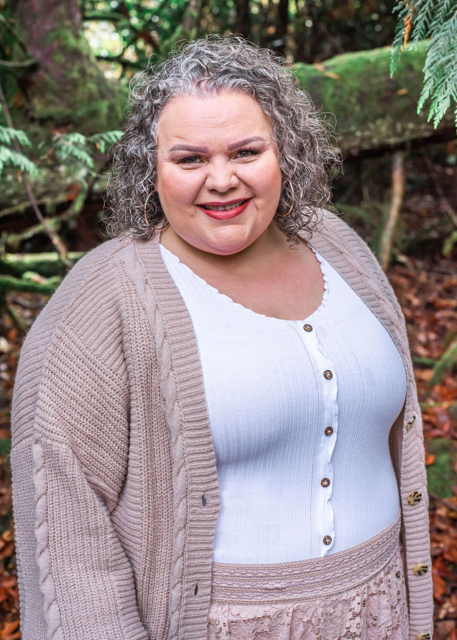 Person in knit cardigan and white top poses against natural forest backdrop.