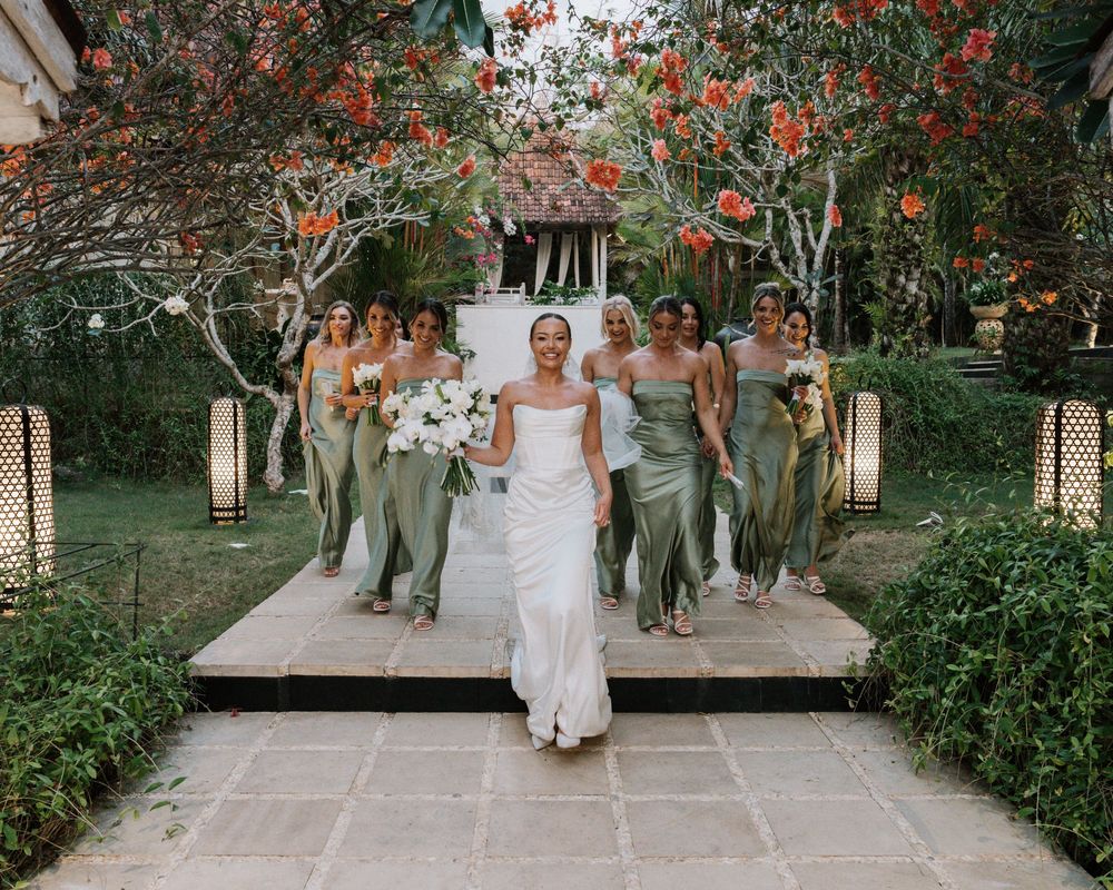 Coral flowers frame a garden pathway as the bridal party in white wedding attire walk through the lush outdoor scene.