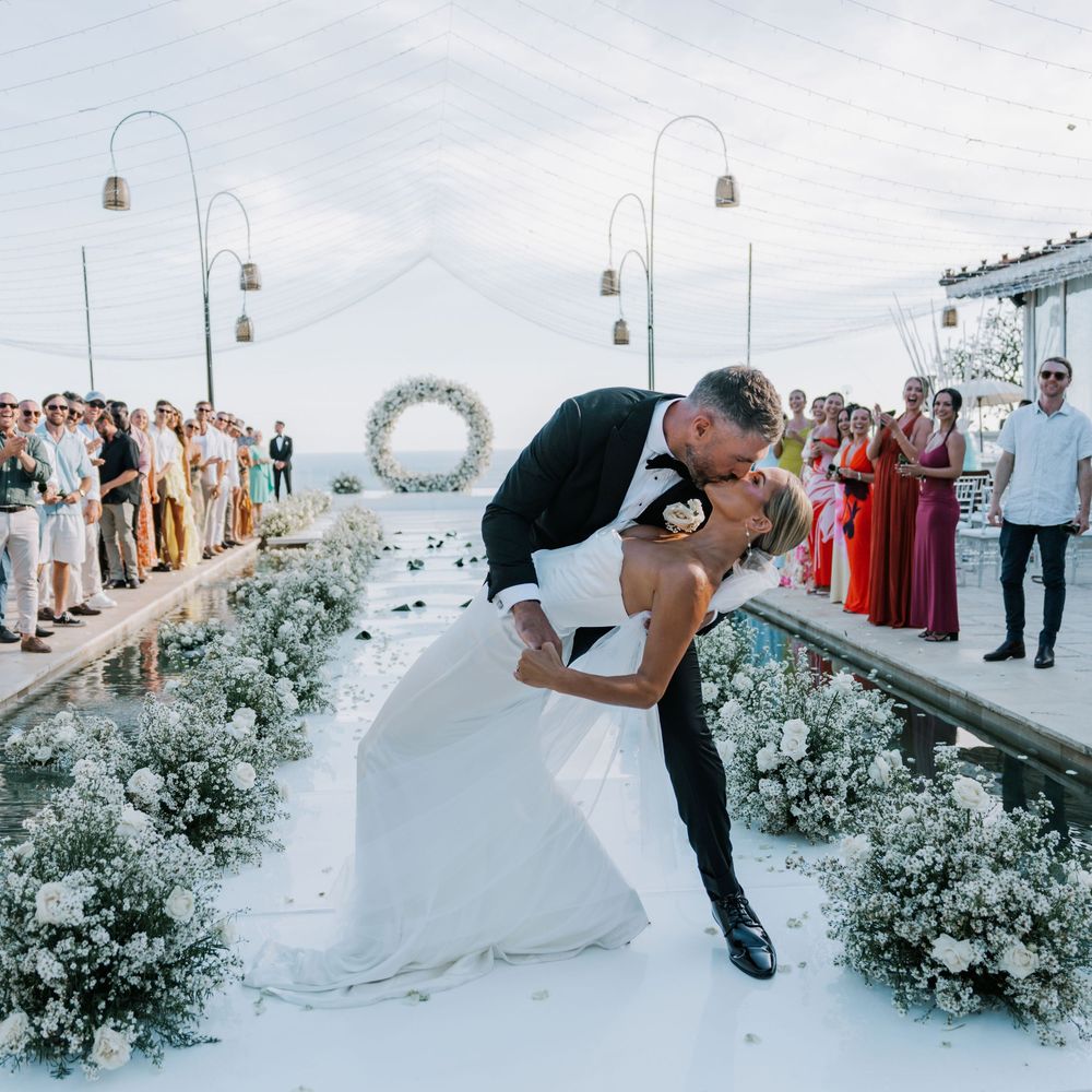 Newlywed couple shares a romantic dip kiss against a backdrop of white floral arrangements and ornate archways during twilight.