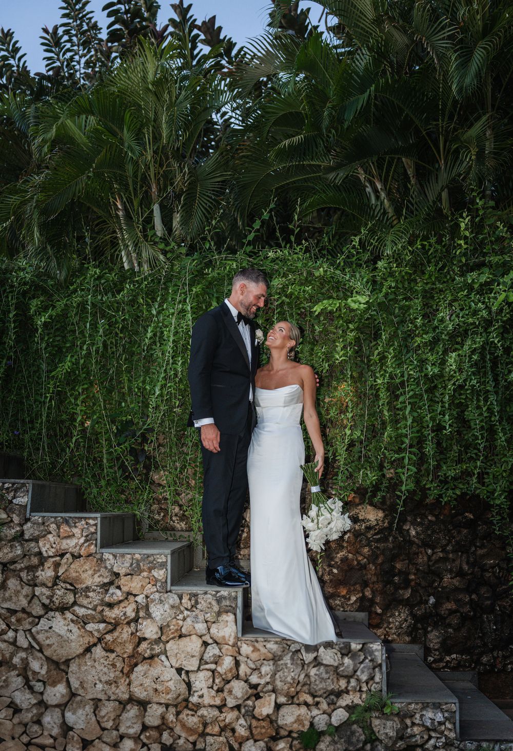 Couple in formal wedding attire share a romantic moment on stone steps surrounded by lush tropical greenery.