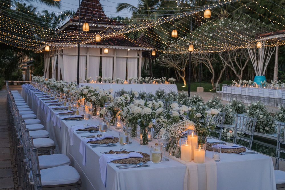 Elegant outdoor wedding reception with glowing string lights, white table settings and floral centerpieces under a clear evening sky.