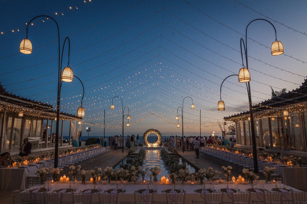 Romantic outdoor dining setup with glowing lanterns and string lights at sunset over the ocean.