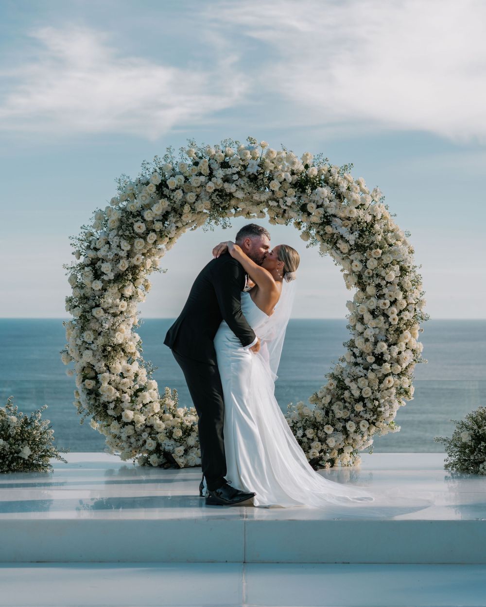 Romantic wedding scene on a balcony by the ocean with a stunning white floral arch and couple embracing under blue skies.