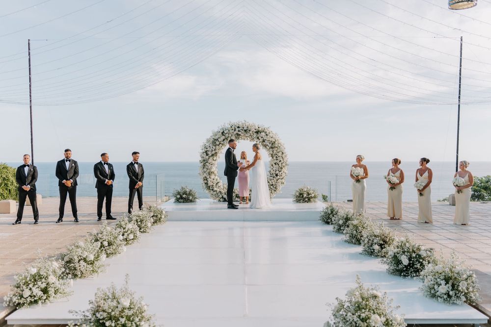 Wedding party gathers at a beachfront ceremony with white floral arch decorations and ocean views.