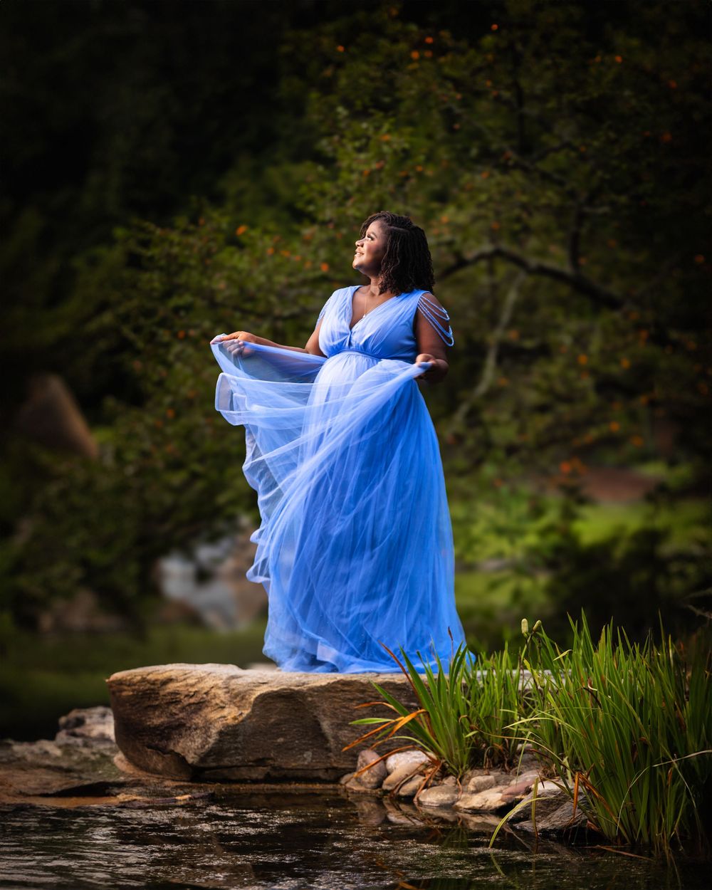 A flowing light blue maternity dress twirls gracefully against a natural outdoor backdrop with rocks and greenery.