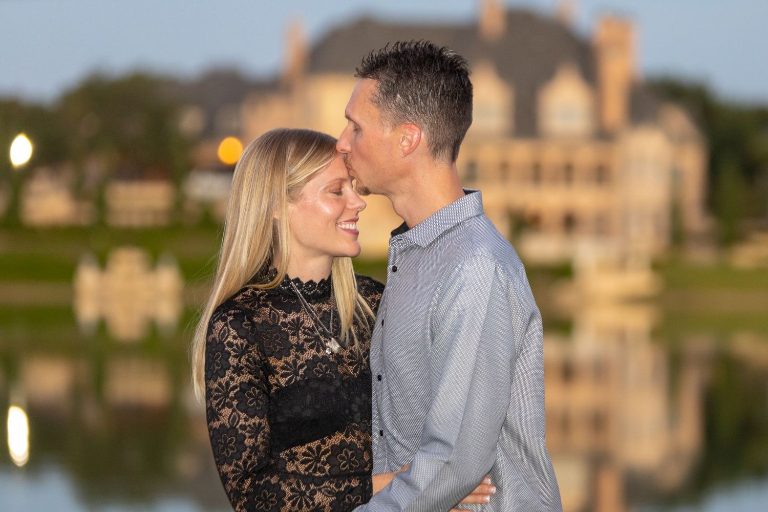 A couple poses together by a serene lake at dusk, with elegant formal attire and a beautiful mansion visible in background.