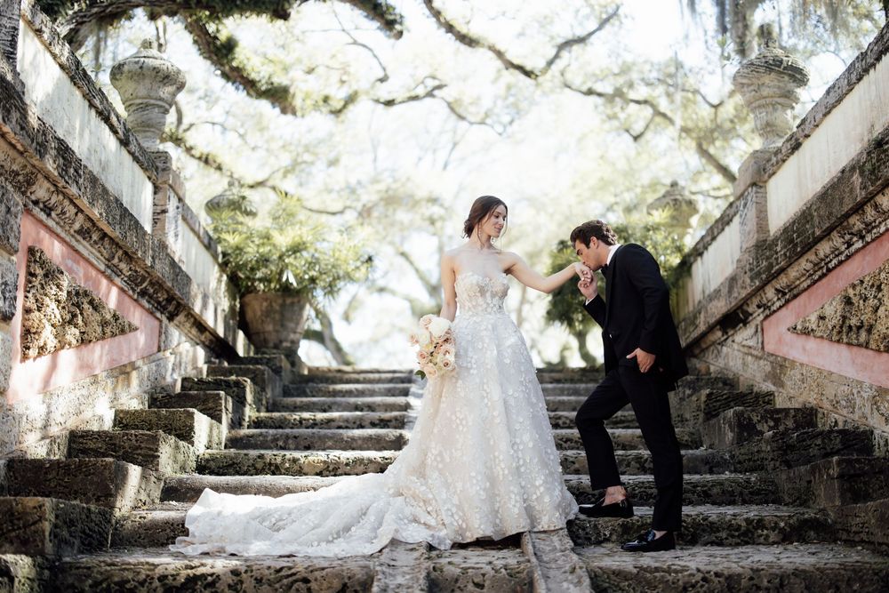 A romantic wedding couple poses on an elegant stone staircase surrounded by overhanging trees and flowers.