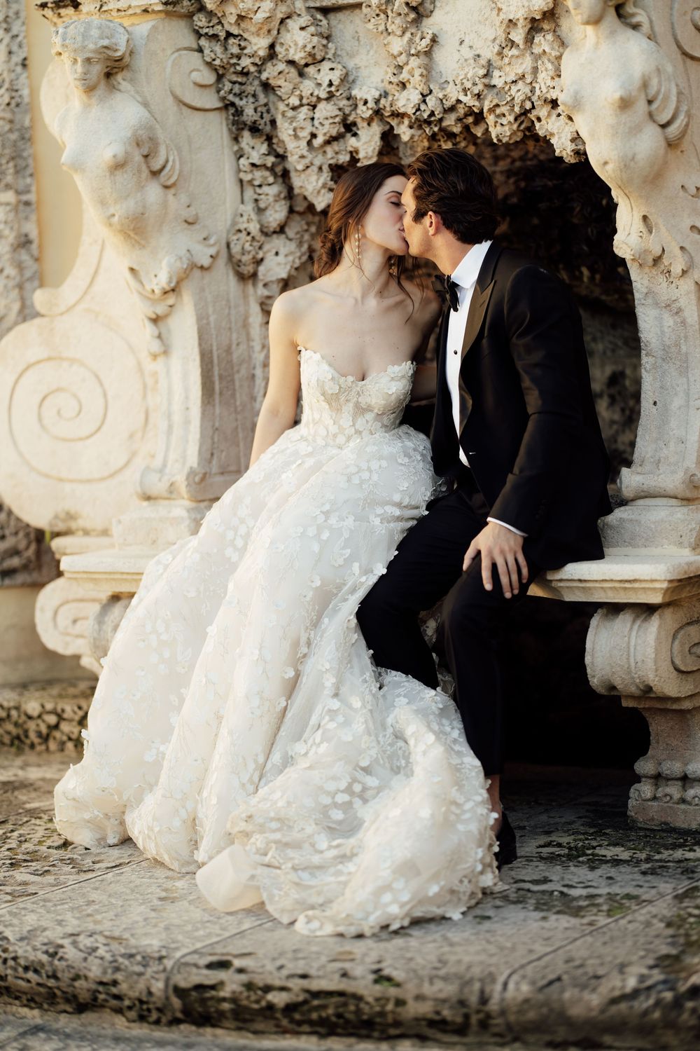 A bride and groom share an intimate moment against an ornate stone wall backdrop with classical architectural details.